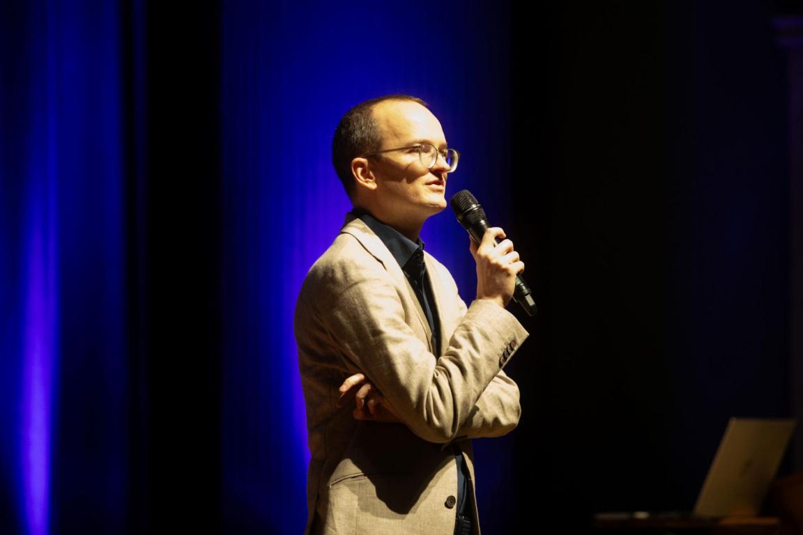 Martin Neukom seitlich vor einem dunklen Hintergrund stehend,  in der Hand ein Mikrophon. 