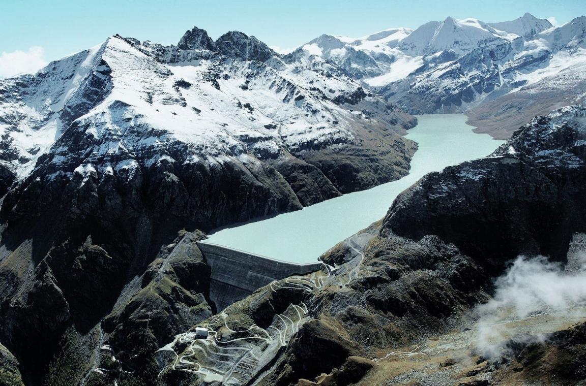 Der Stausee inmitten schneebedeckter Berge ist gut gefüllt. Im Vordergrund sieht man die Staumauer..
