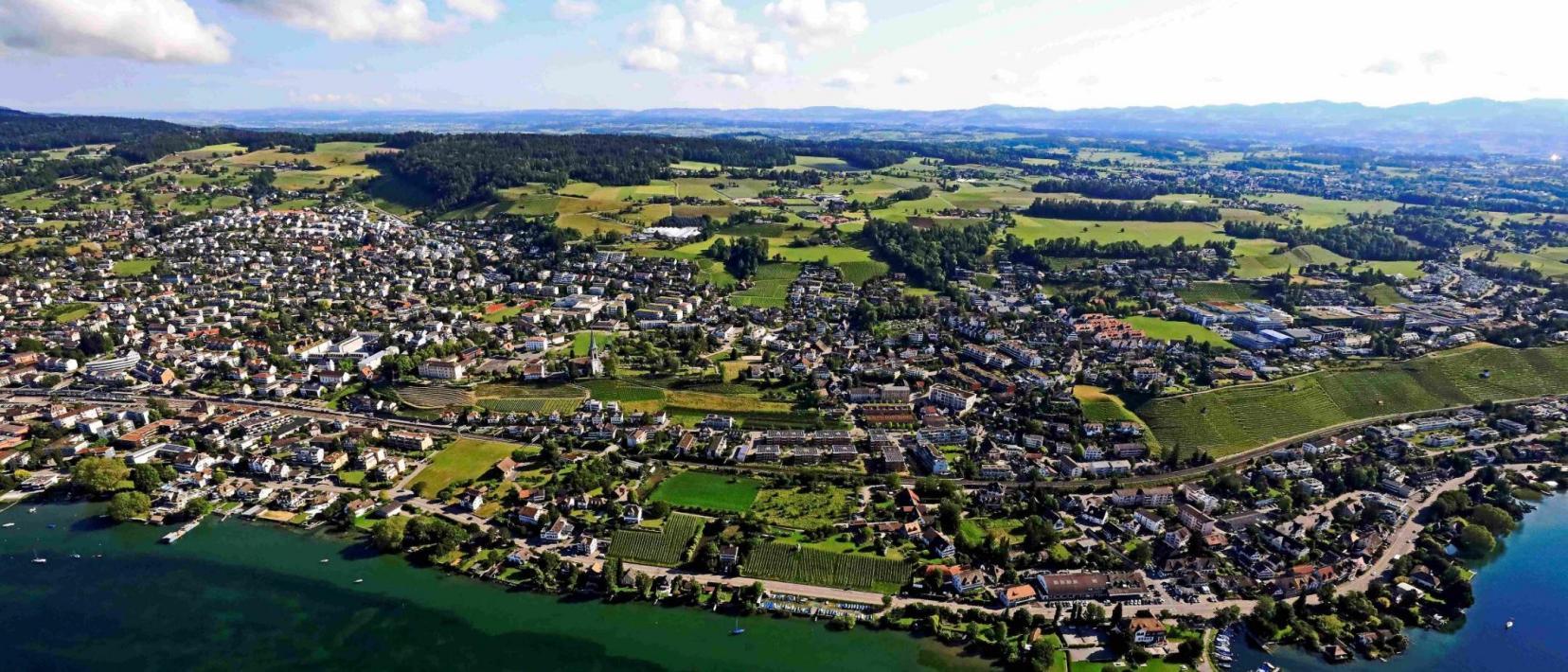 Luftaufnahme der Gemeinde Stäfa mit dem See im Vordergrund und einem blauen Himmel mit Wolken in Hintergrund. 