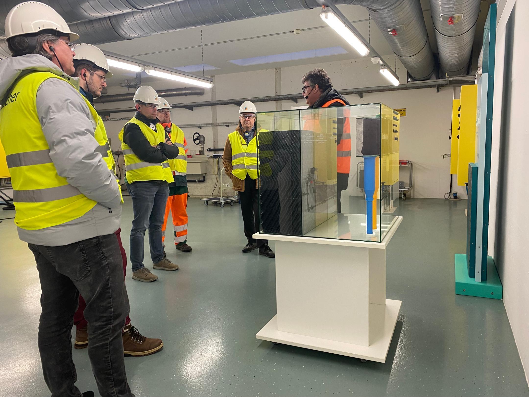 Sechs Männer in Sicherheitswesten und mit Bauhelmen auf dem Kopf stehen um einen Schaukasten aus Glas herum. 