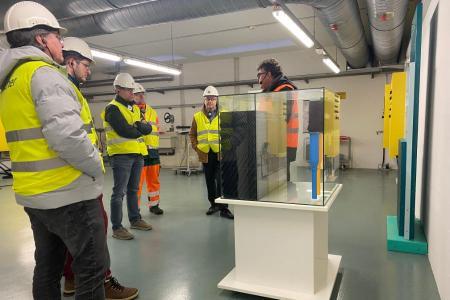 Sechs Männer in Sicherheitswesten und mit Bauhelmen auf dem Kopf stehen um einen Schaukasten aus Glas herum. 