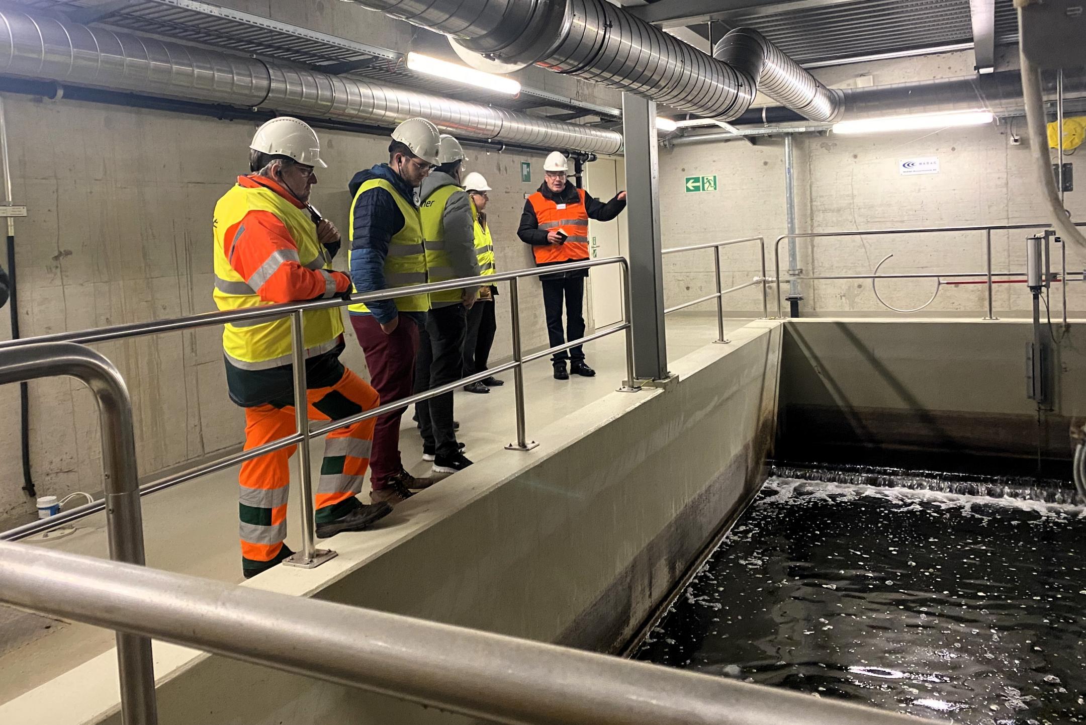 Man sieht vier Personen in Sicherheitswesten und mit weissen Bauhelmen um ein Abwasserklärbecken stehen, während eine Person Erklärungen abgibt. 