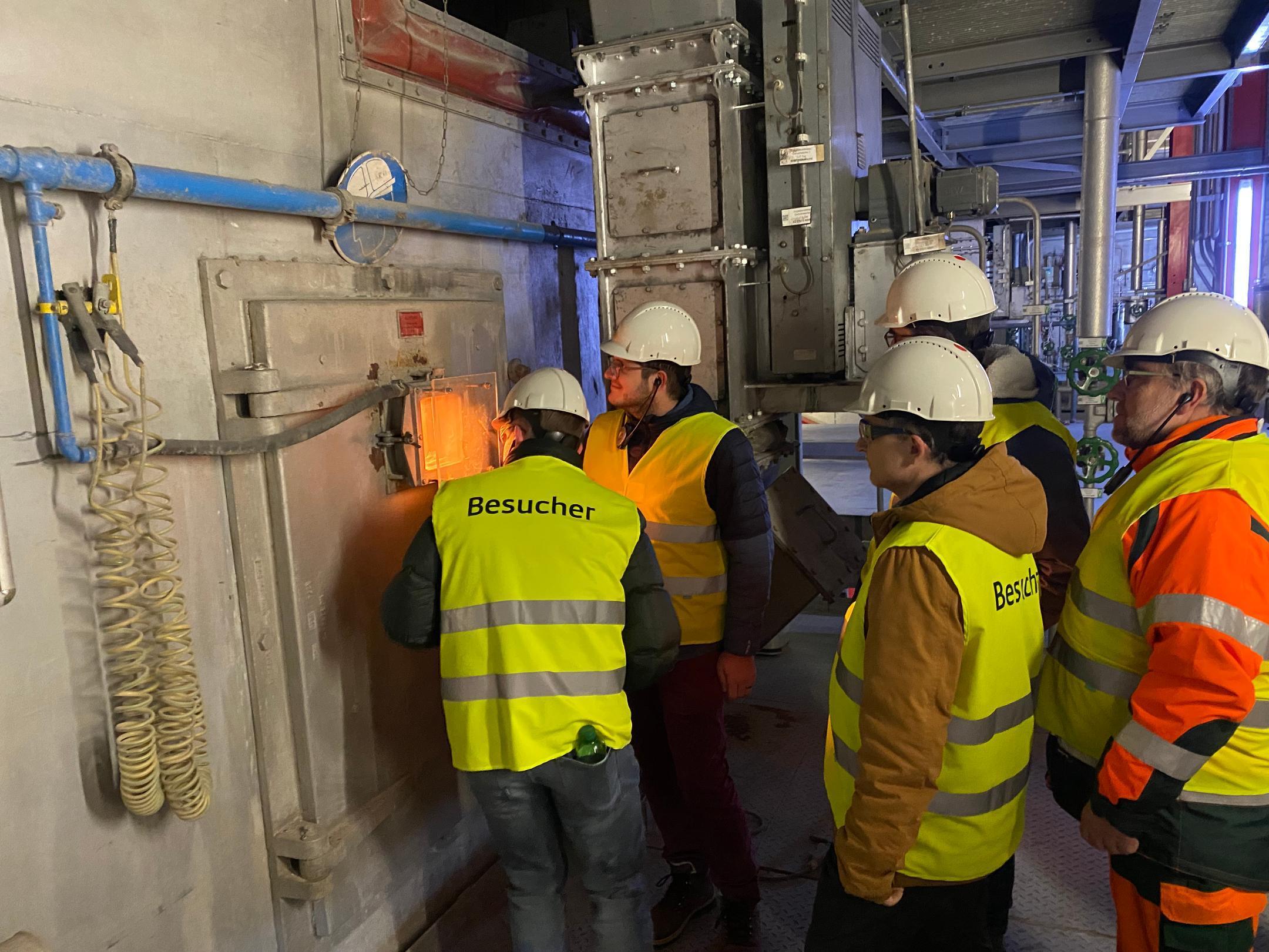 Fünf Personen drängen sich vor der verschlossenen Tür eines Ofens. Nur durch ein kleines Fenster sieht man den orangen Schein des Feuers im Innern. 