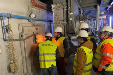 Fünf Personen drängen sich vor der verschlossenen Tür eines Ofens. Nur durch ein kleines Fenster sieht man den orangen Schein des Feuers im Innern. 