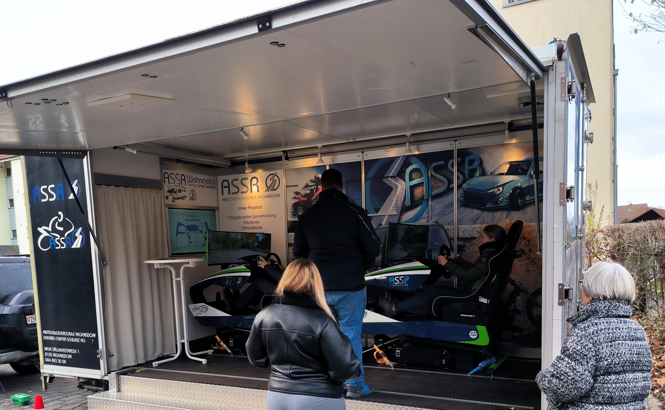 Zwei von hinten fotografierte Frauen sehen zu, wie eine Dame in einem Fahrsimulator sitzt . Ein Instruktor steht neben ihr.