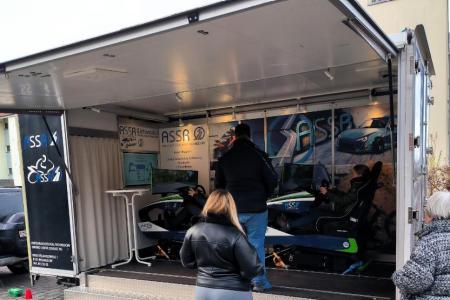 Zwei von hinten fotografierte Frauen sehen zu, wie eine Dame in einem Fahrsimulator sitzt . Ein Instruktor steht neben ihr.