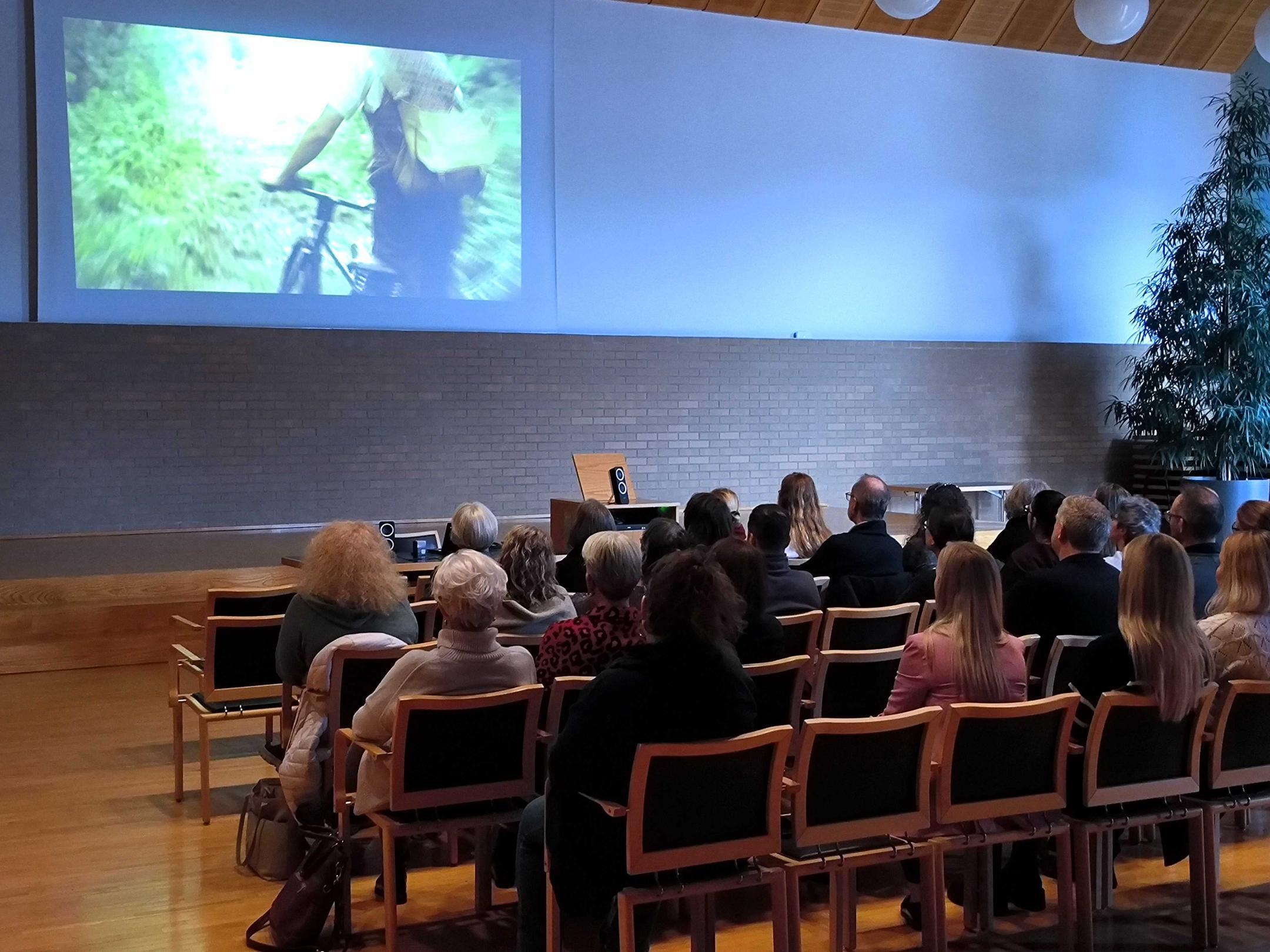 In einem halbdunklen Saal sieht man von hinten rund 30 Personen auf Holzstühlen sitzen. Sie schauen auf eine Leinwand wo das Standbild eines Velofahrers zu sehen ist. 