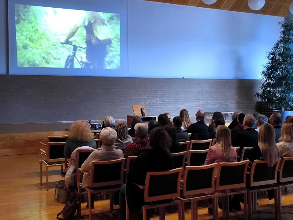 In einem halbdunklen Saal sieht man von hinten rund 30 Personen auf Holzstühlen sitzen. Sie schauen auf eine Leinwand wo das Standbild eines Velofahrers zu sehen ist. 