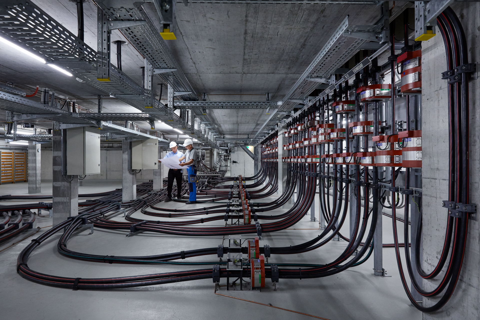 Zwei Männer mit Bauhelm auf den Kopf stehen in einer grossen Betonhalle in der eine Vielzahl elektrischer Installationen zu sehen sind. 