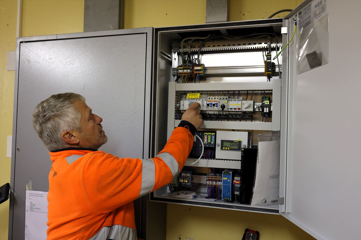 Ein Mann mit grauen Haaren in einer orangen Arbeiterjacke zeigt mit dem Finger auf einen Schalter in einem weit geöffneten silberfarbenen Elektrokasten.