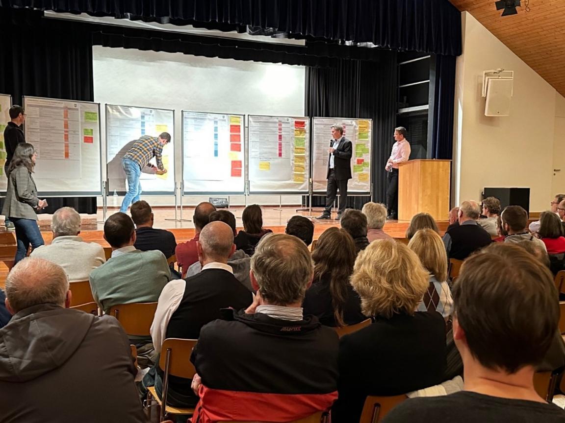 Von hinten sieht man in einem halbdunklen Gemeindesaal das Publikums, während davor auf einer Bühne drei Männer sind. Einer lehnt an ein Stehpult, der zweite hält ein Mikrofon und ein dritter befestigt einen Zettel an einer grossen Tafel.