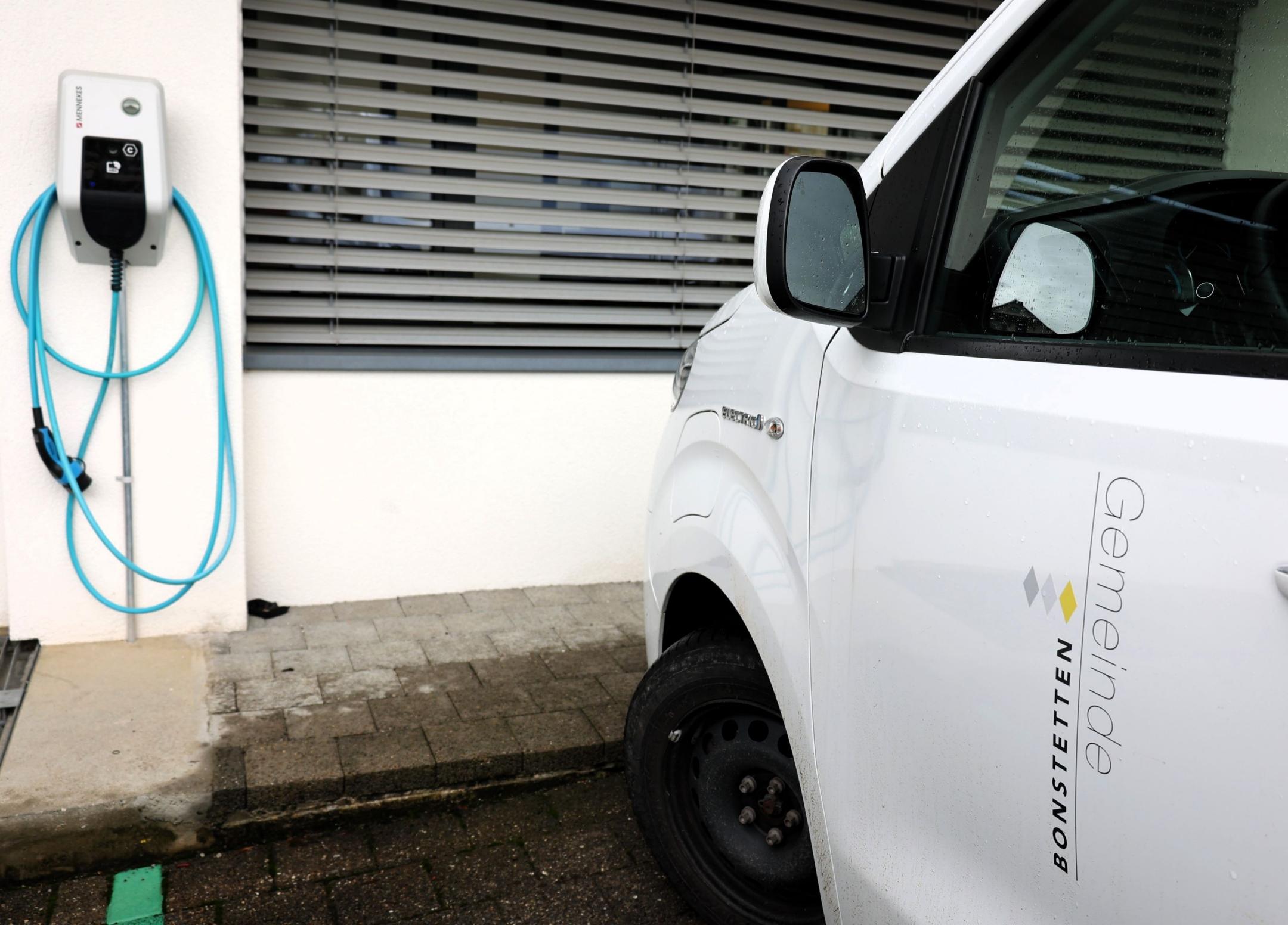 Auf einem Parkplatz steht ein weisses Elektrofahrzeug das auf der rechten Seitentüre die Aufschrift Gemeinde Bonstetten trägt. Dahinter hängt ein Stromladegerät an der Wand.
