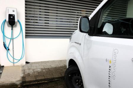 Auf einem Parkplatz steht ein weisses Elektrofahrzeug das auf der rechten Seitentüre die Aufschrift Gemeinde Bonstetten trägt. Dahinter hängt ein Stromladegerät an der Wand.