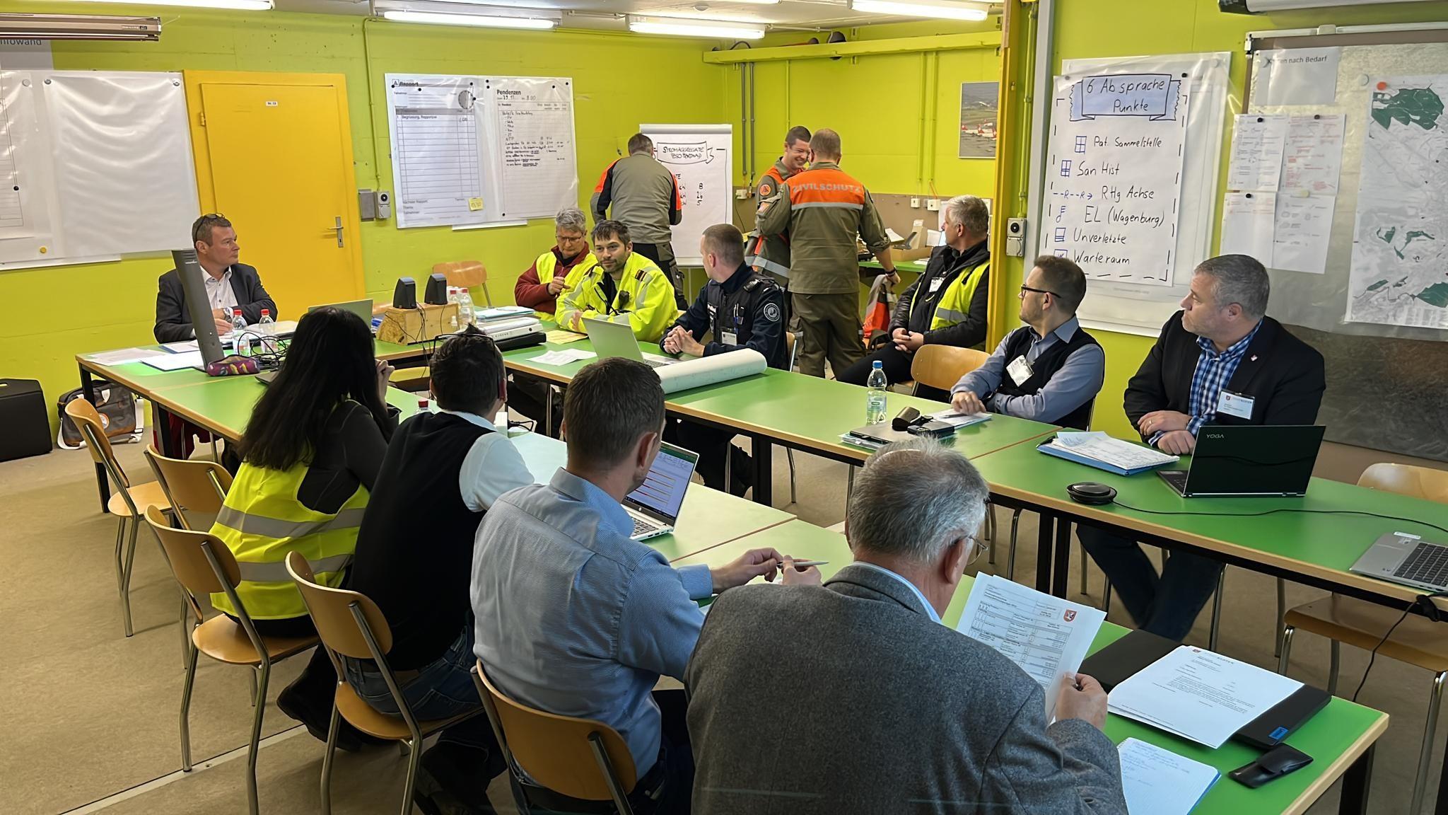 In einem hellbeleuchteten Raum sitzt knapp ein Dutzend Personen an Tischen mit grüner Tischplatte beim Rapport. 