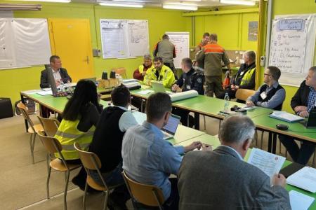 In einem hellbeleuchteten Raum sitzt knapp ein Dutzend Personen an Tischen mit grüner Tischplatte beim Rapport. 