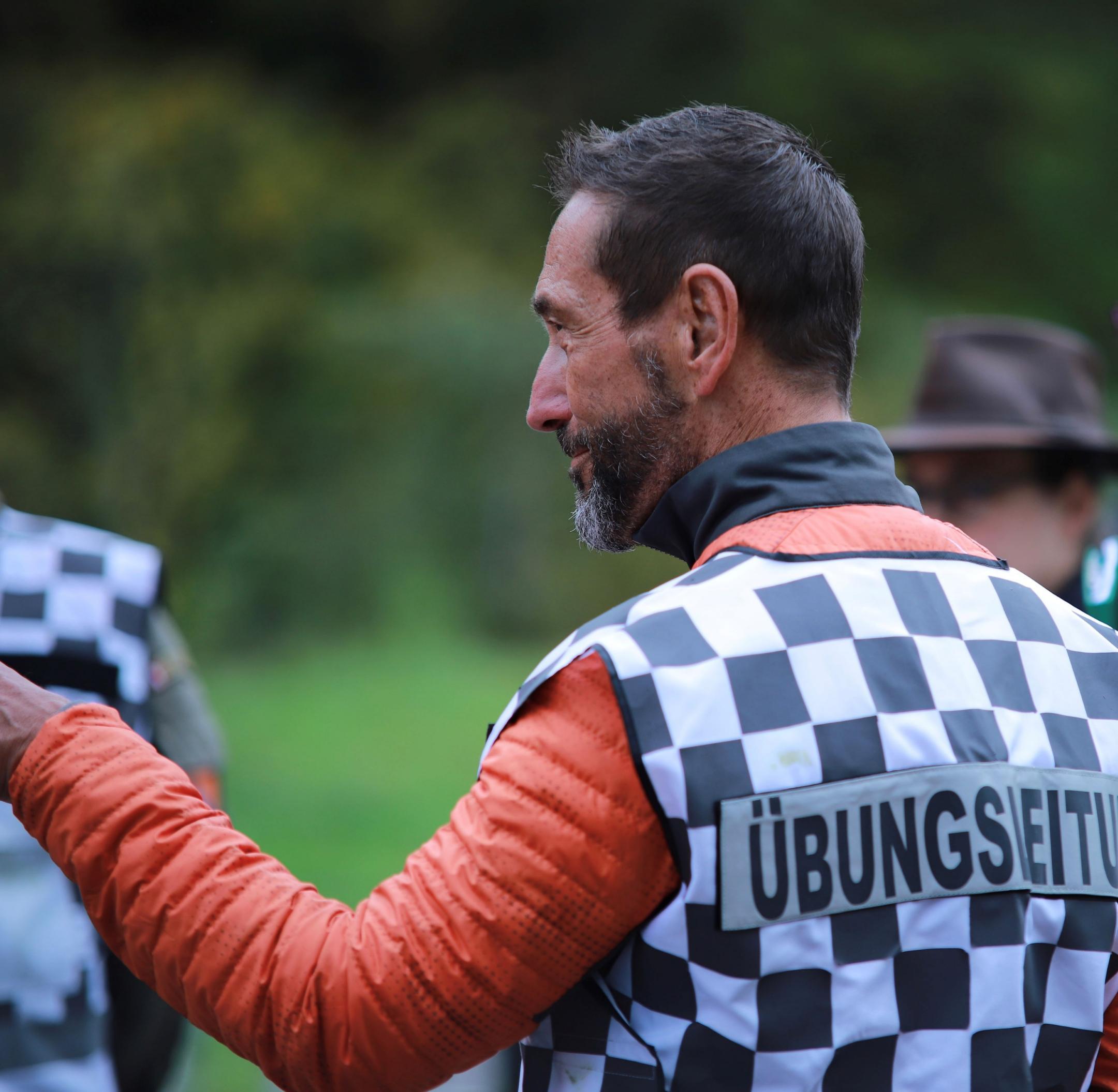 Markus Meile in einer Profilansicht. Er trägt eine rote Jacke und eine schwarz-weisse Weste mit der Aufschrift Übungsleiter. 