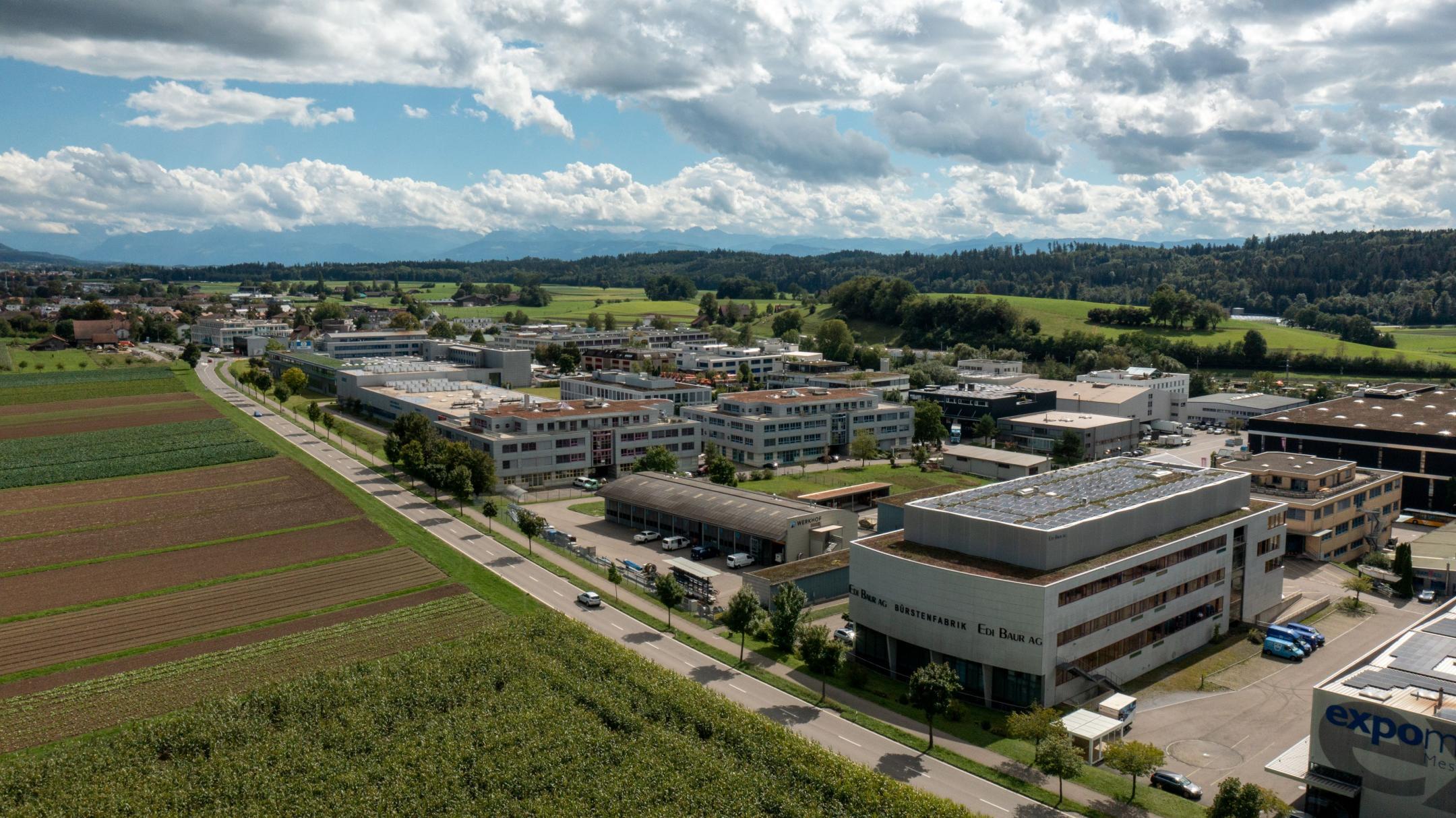 Eine Luftaufnahmen des Industriegebiets der Gemeinde Fehraltorf.. Es ist eingebettet in eine grüne Umgebung, im Vordergrund sind Ackerflächen, am Horizont ein bewaldeter Hügelzug.