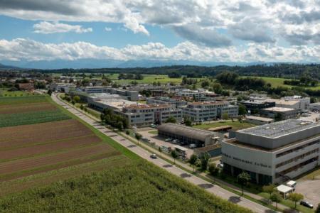 Eine Luftaufnahmen des Industriegebiets der Gemeinde Fehraltorf.. Es ist eingebettet in eine grüne Umgebung, im Vordergrund sind Ackerflächen, am Horizont ein bewaldeter Hügelzug.