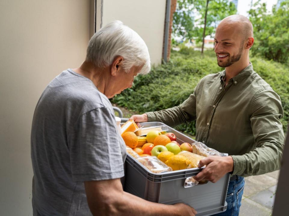 Ein jüngerer Mann bringt einer Senioren Lebensmittel in einer offenen Transportbox an die Haustüre. 