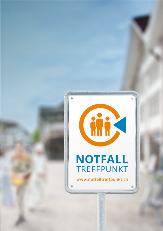 Vor einem verschwommenen Hintergrund, auf dem noch einige Menschen und Häuser erkennbar sind, steht ein Schild mit der Aufschrift Notfalltreffpunkt.