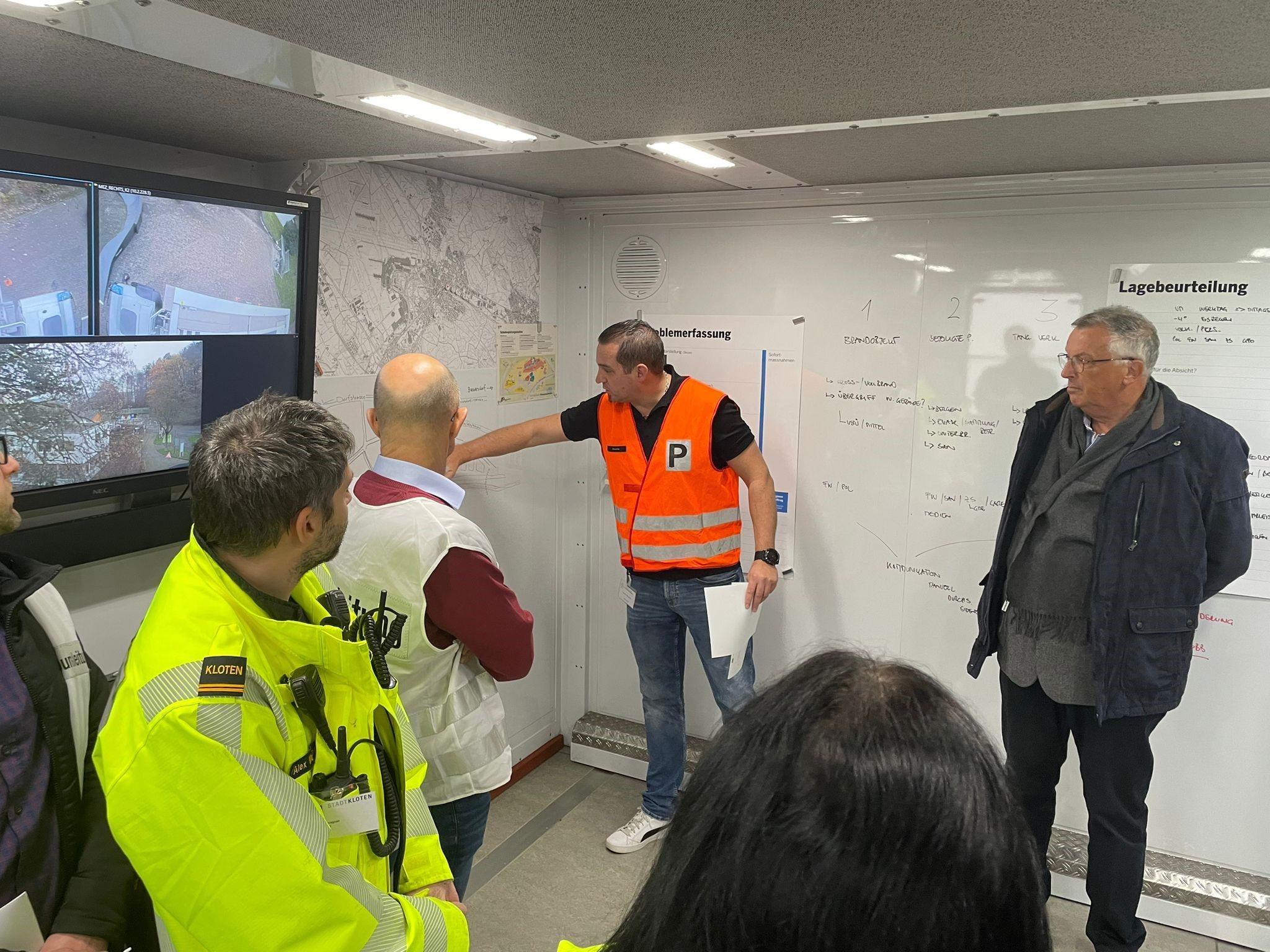 In einem provisorisch eingerichteten Kommandoposten mit vielen Plänen und Notizen an den Wänden halten mehrere Personen einen Lagebesprechung.