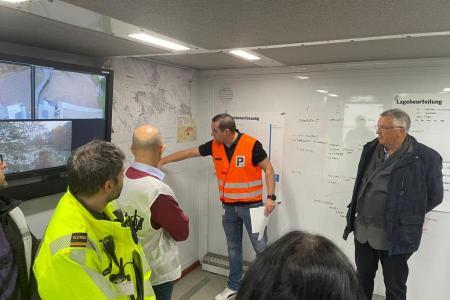 In einem provisorisch eingerichteten Kommandoposten mit vielen Plänen und Notizen an den Wänden halten mehrere Personen einen Lagebesprechung.