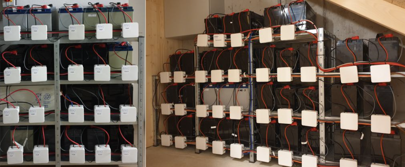 Zwei Regalwände mit dutzenden gebrauchten Batterieblöcken in einem Kellerraum, jeder Block über ein weisses Modul und rote sowie schwarze Kabel verbunden.