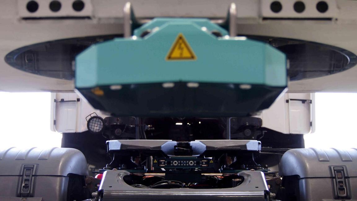 Symmetrische Untersicht des Testfahrzeugs: Hochvolt-Bauteil mit Warnsymbol im Zentrum, darunter die Sattelkupplung, seitlich die Reifen von Zugmaschine und Auflieger.
