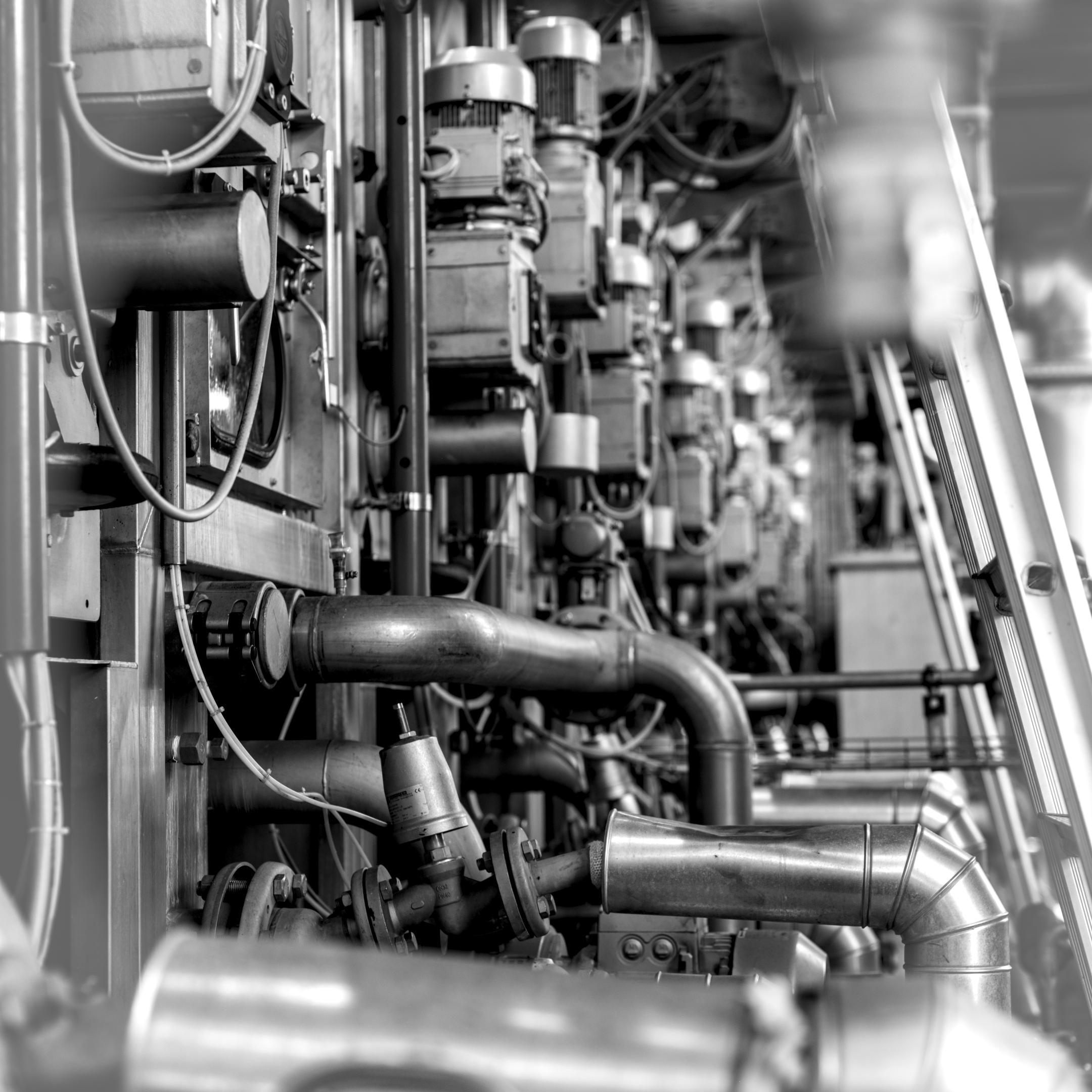 Schwarz-Weiss-Aufnahme eines Industrieanlagen-Innenraums mit Rohrleitungen, Ventilen, Elektromotoren und Kabeln.