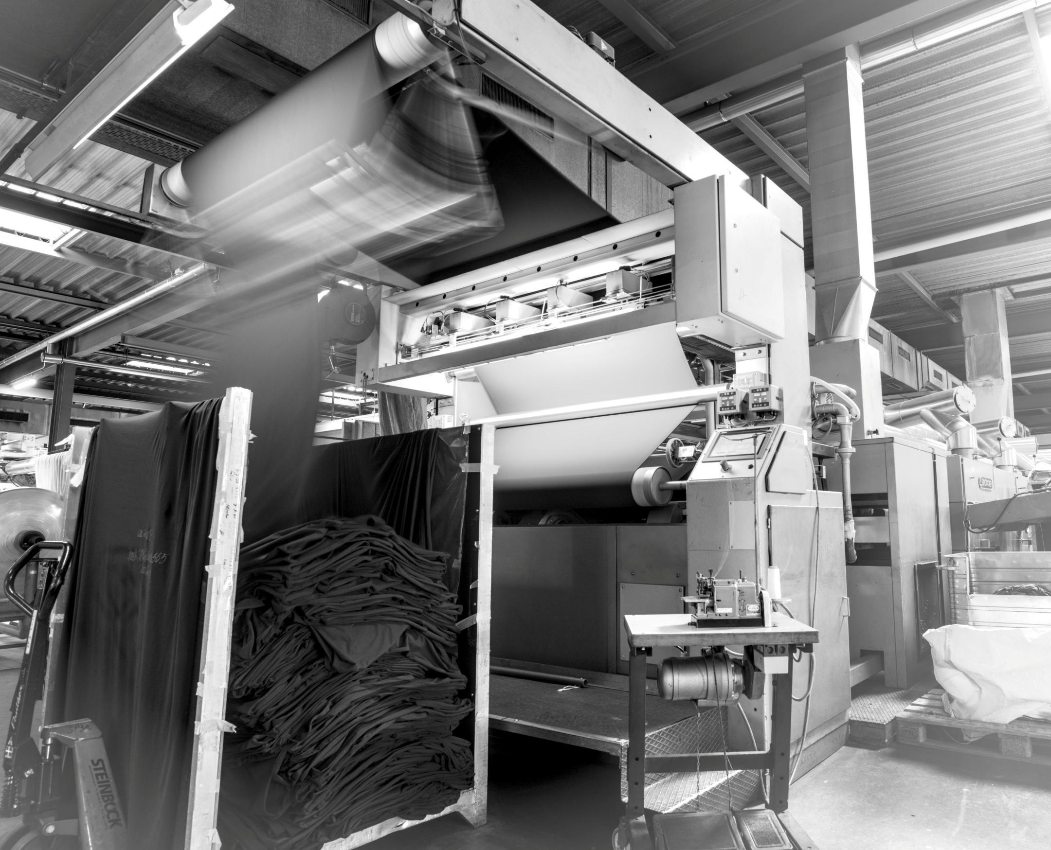 Schwarz-weisses Bild einer Textilmaschine, die dampfbetrieben schwarzen Stoff fixiert, mit Stapeln des gefalteten Materials und der Aufschrift «STEINBOCK» auf einer Palette.