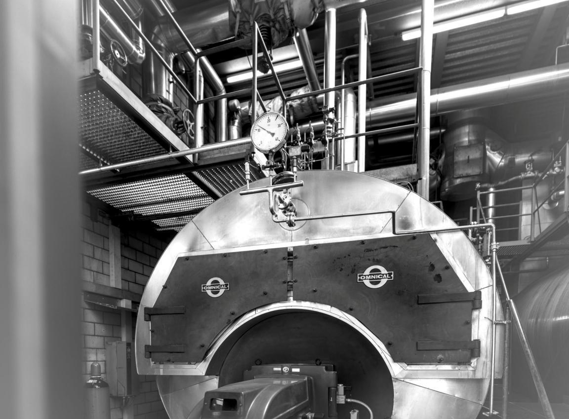 Grossaufnahme eines Dampfkessels mit Manometer und Brenner in einer Industriehalle, im Hintergrund isolierte Dampfleitungen und ein Gitterrostpodest.