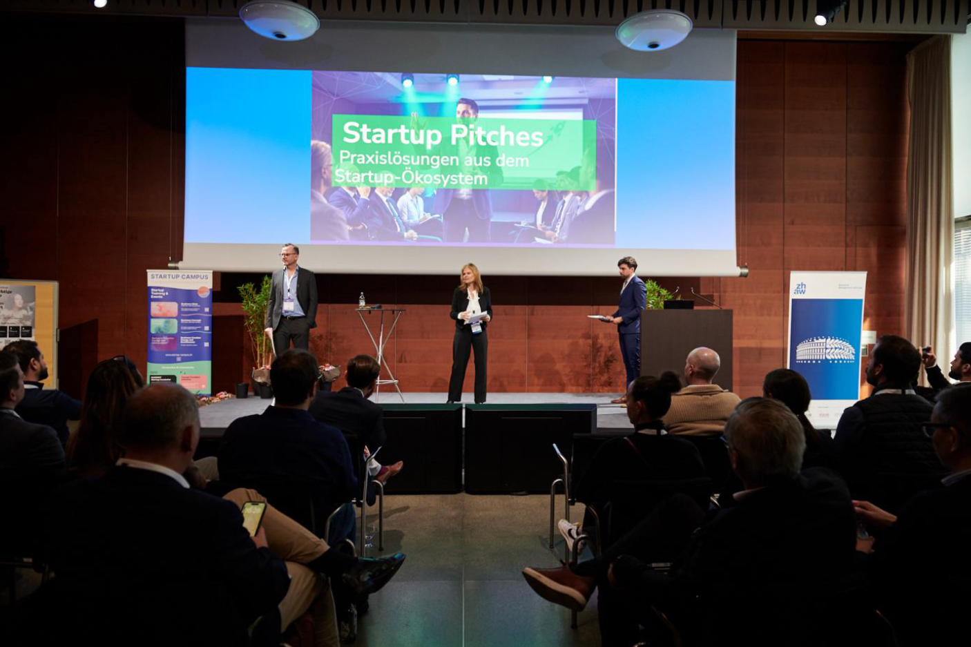 Auf einer Bühne moderieren drei Personen die «Startup Pitches», im Hintergrund eine Leinwand, davor ein Publikum im Saal.