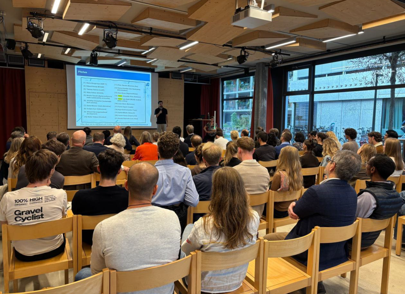 Ein Redner präsentiert «Pitches» auf einer Leinwand vor einem Publikum von rund 50 Personen in einem Vortragssaal.