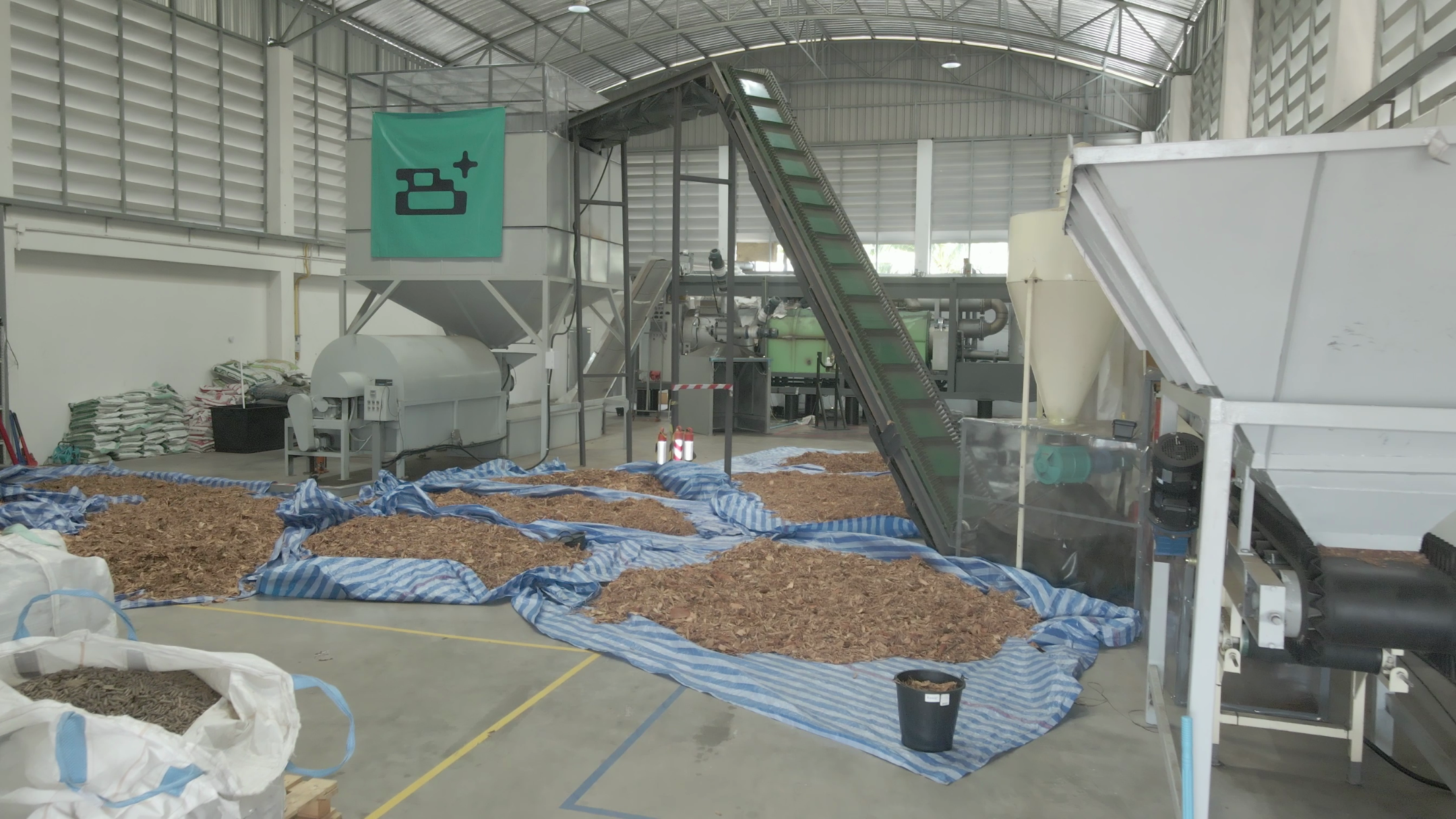 Pilotfabrik in Thailand mit Schreddergut auf blauen Planen und einer Förderanlage, im Hintergrund Maschinen und ein grünes Logo mit symbolisiertem Akku.