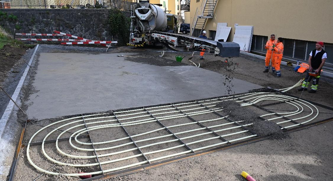 Bauarbeiter bringen mit einem Förderband Zementmörtel auf vorbereitete Kunststoffrohre für eine Flächenheizung auf.
