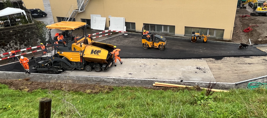 Mitarbeiter in orangefarbener Arbeitskleidung bei der Asphaltierung einer Fläche mit einem gelben Asphaltfertiger und zwei Walzen. Im Vordergrund liegen orange Kunststoffrohre, um Überhitzung zu vermeiden. Der Fertiger trägt die Aufschrift «KE».