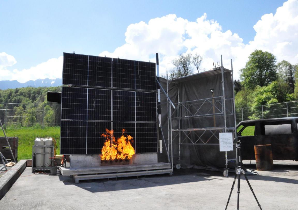 Experimenteller Brandversuch im Freien: Ein offenes Feuer brennt vor einer Wand aus Photovoltaik-Modulen.