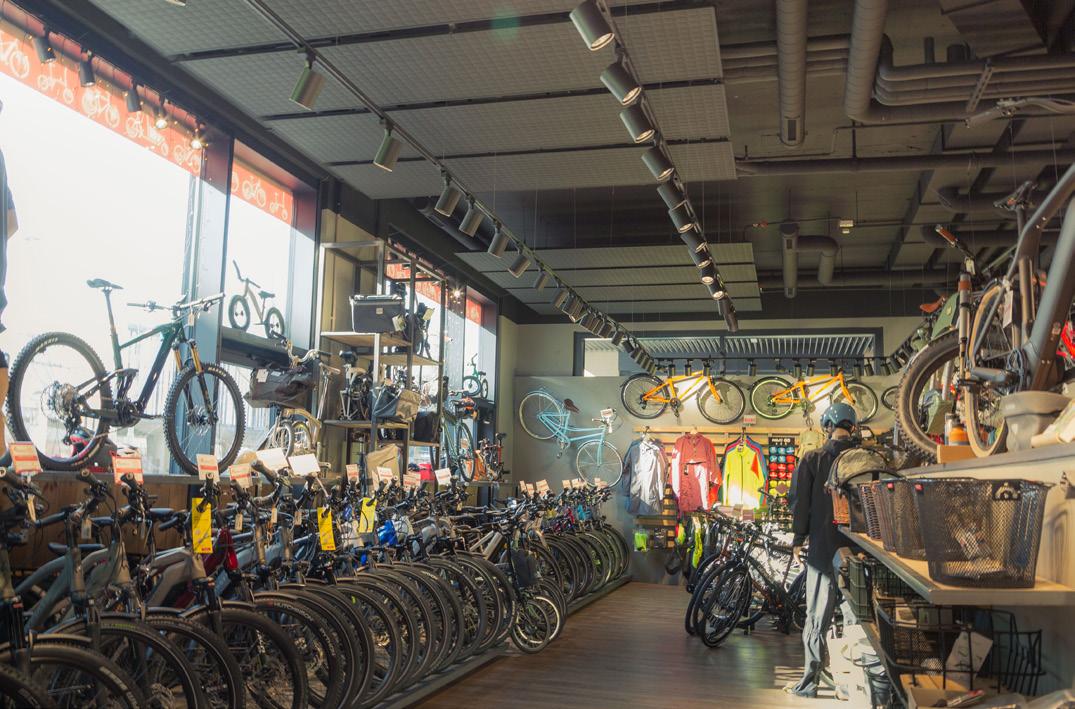 Innenansicht eines Fahrradgeschäfts mit Reihen von Fahrrädern, Bekleidung und Zubehör unter einer Decke mit vielen Leuchten.