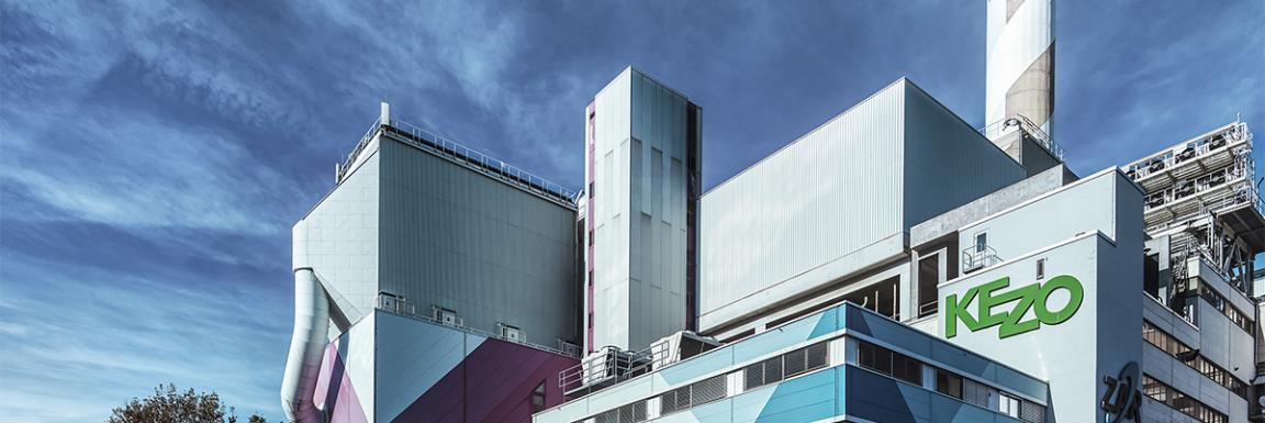 Industriegebäude der KEZO mit markanter Fassade, Schornstein und klarer blauer Himmel im Hintergrund.