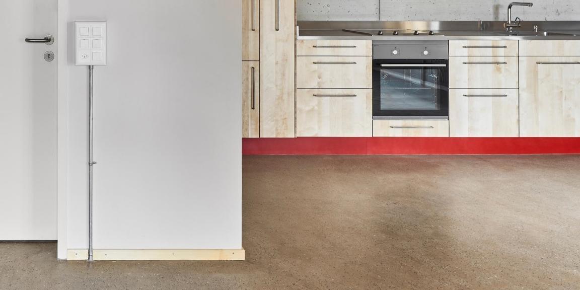 Moderne Küche mit hellen Holzfronten und roter Sockelleiste, im Vordergrund ein glatter Terrazzo-Boden.