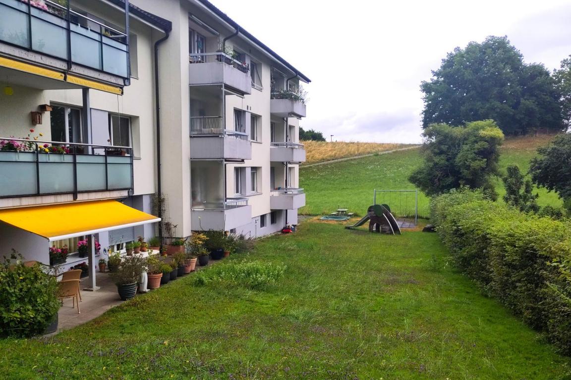 Gartenansicht einer Wohnsiedlung mit Balkonen, darunter eine Terrasse mit gelber Markise, und einer grossen Rasenfläche mit Spielplatz und Hügel im Hintergrund.