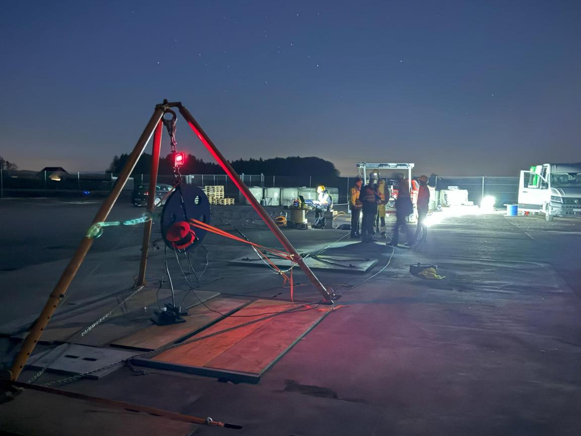 Aufnahme einer nächtlichen Messkampagne auf einer befestigten Fläche. Ein Dreibein-Stativ mit Messgerät steht im Vordergrund, im Hintergrund sind mehrere Personen und ein Fahrzeug mit Beleuchtung sichtbar.
