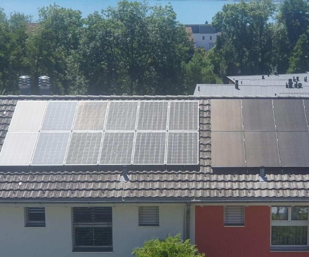 Dach eines Wohnhauses mit zwei Reihen Photovoltaikmodulen. Links mehrere helle Module, rechts dunkle Module. Im Hintergrund grüne Bäume und Dächer.