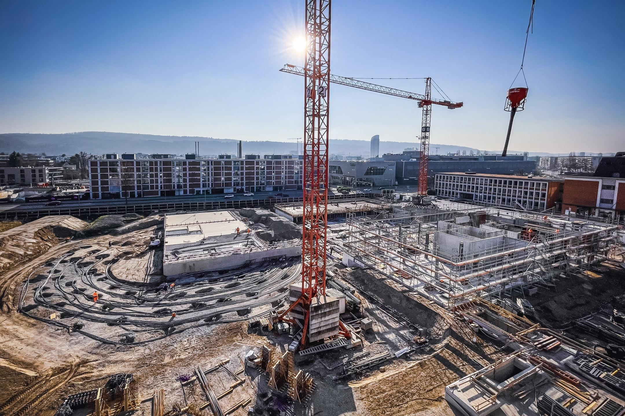 Baustelle mit zwei Baukränen, konzentrischen Rohrleitungen für das Erdsondenfeld und Gebäuden im Hintergrund bei sonnigem Wetter.