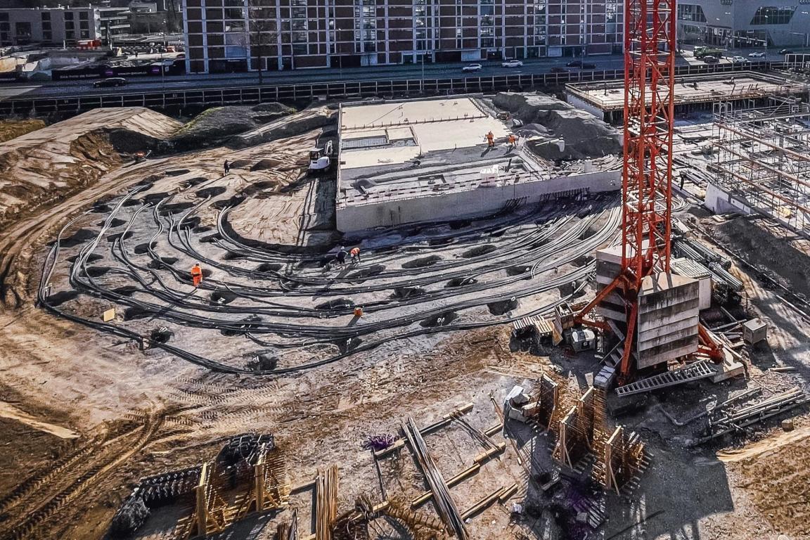 Luftaufnahme einer Baustelle mit konzentrisch angeordneten Rohrleitungen für ein Erdsondenfeld, Baukran und Gebäudestrukturen im Hintergrund