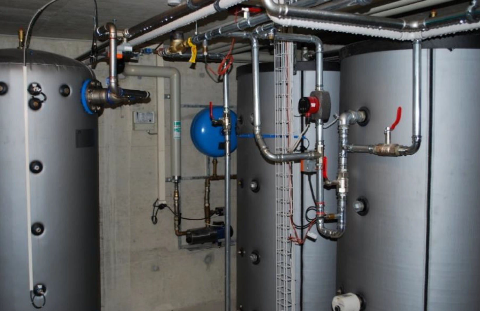 Einer der beiden Verdampferspeicher (links) und die beiden Heizungsspeicher. Im Hintergrund sind der Druckbehälter (blau) und darunter der Kompressor der Regenwasseranlage sichtbar. Zum Zeitpunkt der Aufnahme sind die Leitungen noch nicht isoliert.