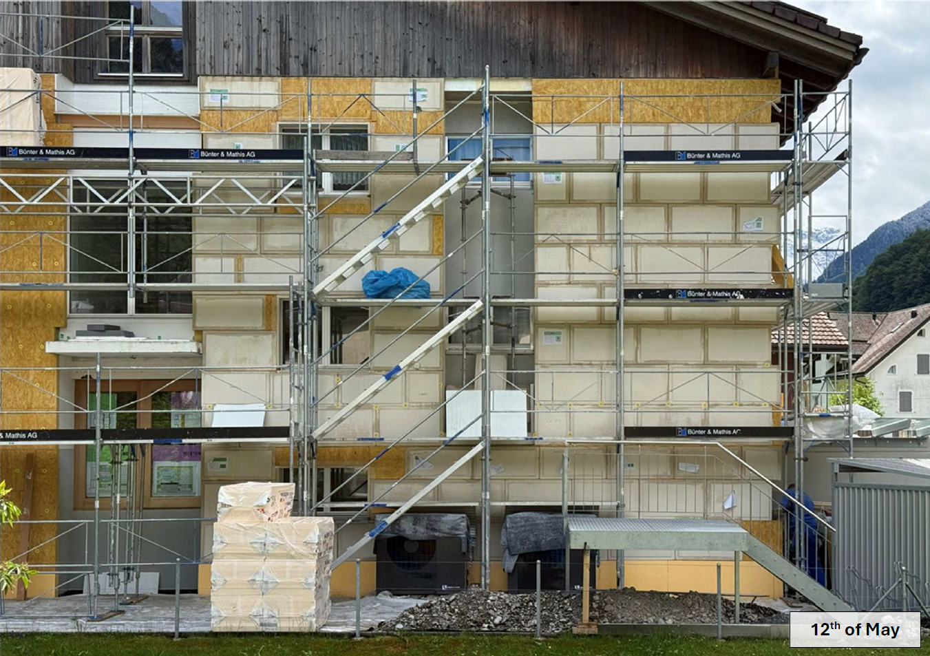 Eingerüstetes Mehrfamilienhaus in Sanierung, an der Fassade weisse vorgefertigte Dämmpaneele mit Etiketten und daneben gelbliche Holzwerkstoffplatten mit Mineralwolldämmung.