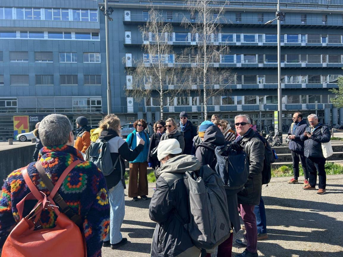 Das Bild zeigt eine Gruppe von Menschen im öffentlichen Raum. Eine Person erklärt etwas, die anderen hören ihr zu. 