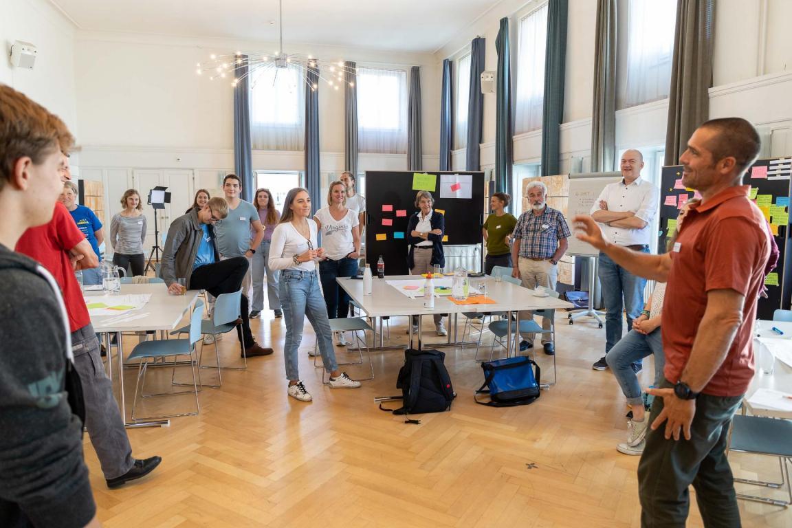 Das Bild zeigt eine Workshop-Situation. Man sieht eine Gruppe von Menschen. Sie stehen in einem grösseren Raum. Eine Person im Vordergrund erklärt den anderen etwas. Im Raum stehen auch Pinnwände mit angepinnten Moderationskarten. Ebenfalls gibt es Tische, an denen die anwesenden Personen zusammen gearbeitet haben. 