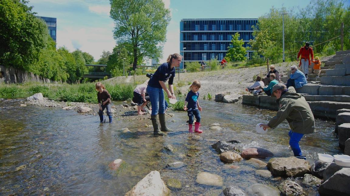 Ein Abschnitt eines Bachs, der revitalisiert ist. Erwachsene und Kinder sitzen auf grossen Steintreppen am Fluss. einige stehen mit Gummistiefeln im untiefen Wasser. 